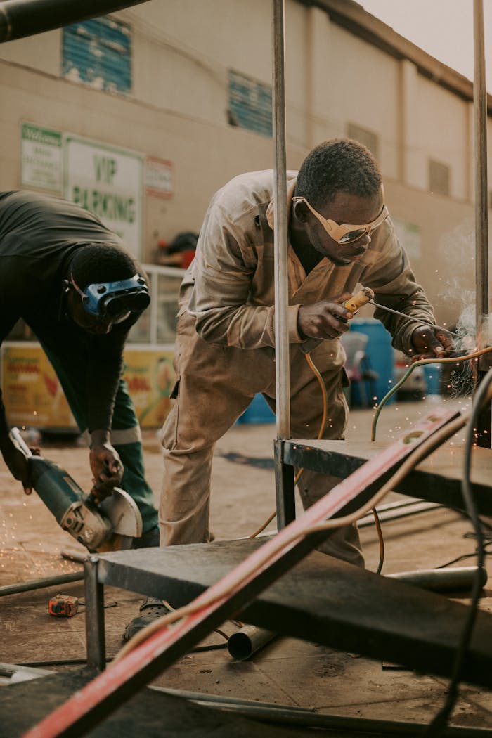 Home Two male workers wearing safety gear welding metal outdoors, showcasing teamwork and industrial labor.